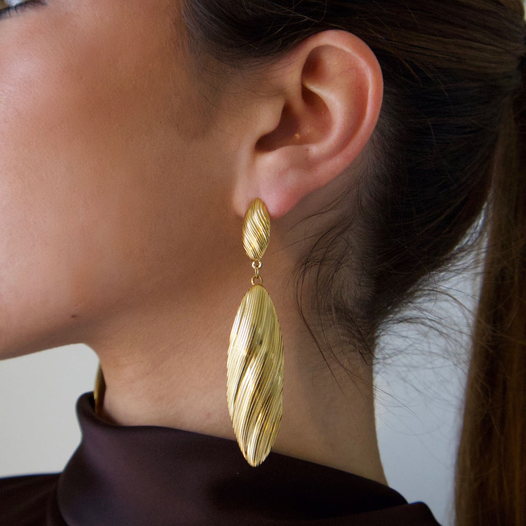 Long gold drop earrings worn on ear with twisted textured elongated design
